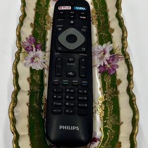 May include: A black remote control with the words "NETFLIX" and "VUDU" on the top. The remote is sitting in a white and green porcelain dish with gold trim and floral designs.