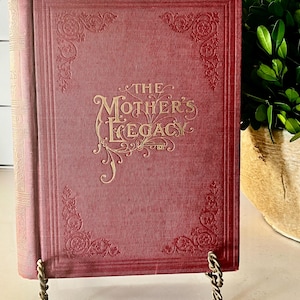 May include: A vintage book titled "The Mother's Legacy" in a deep red hue. The cover features ornate gold lettering and decorative corner designs. The book is displayed on a decorative metal stand, showcasing its aged aesthetic.