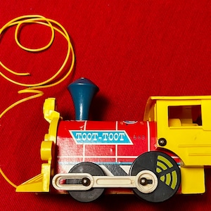 May include: A red, yellow, and blue Toot-Toot train with a yellow caboose and a blue smokestack. The train has a pull string and wheels that turn. The train is on a red surface.