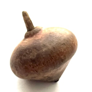 May include: A brown, rounded, hand-carved wooden top with a small, cylindrical stem. The top has a smooth, textured surface and a slightly irregular shape. The color is a mix of light and dark browns.