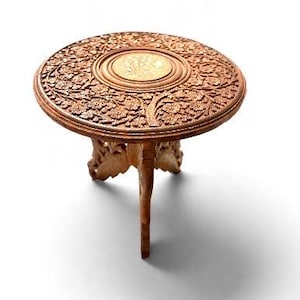 May include: A small, round, brown wooden table featuring intricate floral carvings across the surface and a central circular inlay. The table is supported by three carved legs that extend from the center, showcasing detailed craftsmanship.