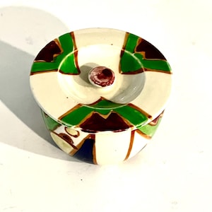 May include: A small, round ceramic trinket box with a lid. The box is cream-colored with green, brown, and gold geometric designs. The lid has a small, round knob. The box is likely used for storing small items like jewelry or keepsakes.