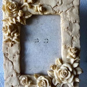 May include: Cream-colored rectangular picture frame with a textured, cracked appearance. The frame is adorned with sculpted roses at the corners and along the sides. The frame has a neutral color and a vintage aesthetic.