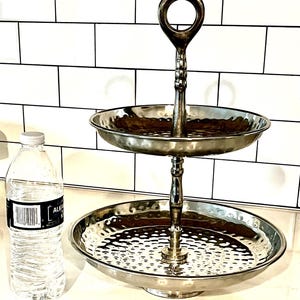 May include: A two-tiered silver serving tray with a hammered texture on the plates and a handle at the top. The tray is designed for displaying food or decorative items. A clear water bottle is next to the tray.