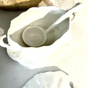 May include: A white porcelain gravy boat with a matching ladle. The boat has a scalloped edge and a handle. The ladle rests inside the boat. A white saucer is beneath the boat. The items are on a light-colored surface.