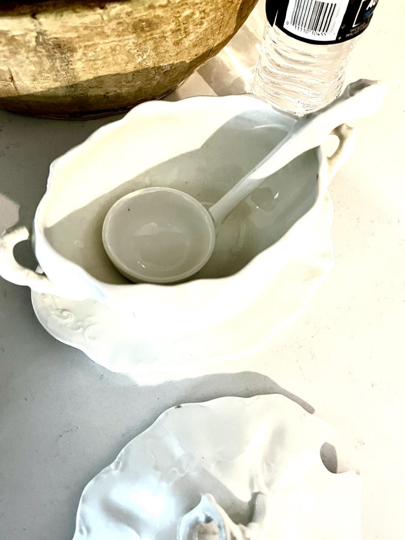 May include: A white porcelain gravy boat with a matching ladle. The boat has a scalloped edge and a handle. The ladle rests inside the boat. A white saucer is beneath the boat. The items are on a light-colored surface.