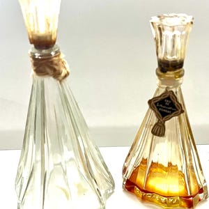 May include: Two vintage perfume bottles. One is clear with a textured, flared design, and the other is amber-colored with a label that reads "Eau de Bombay". Both have decorative stoppers and are displayed against a white background.