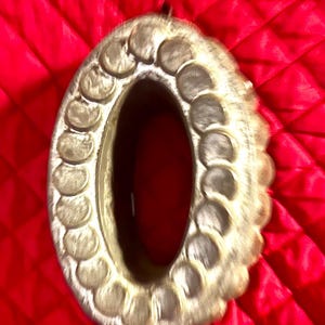 May include: An antique, oval-shaped, silver-toned metal frame with a scalloped edge. The frame has a dark interior and a small loop at the top for hanging. The background is a red, quilted fabric.