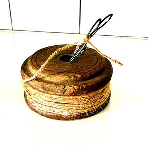 May include: A wooden spool with a dark brown finish, wrapped with natural twine. A metal clip holds the twine in place. The spool has a hole in the center and is set against a white background.