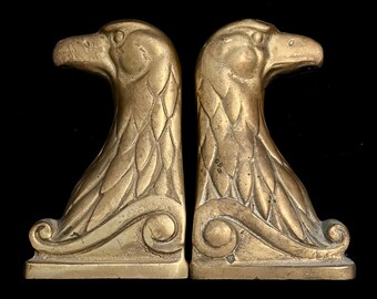 May include: A pair of gold-toned metal bookends shaped like eagles with their heads turned to the side. The eagles are perched on a decorative base with a scroll design.