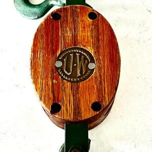 May include: A vintage wooden pulley with a metal hook and bracket. The pulley has a brown wooden body with a brass emblem that reads "U-W". The metal components are painted green.