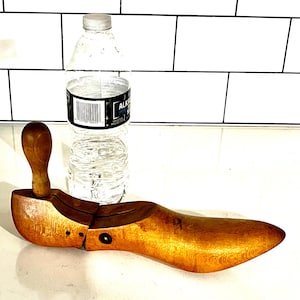 May include: A wooden shoe last, a form used to shape shoes, is displayed. The last is a light brown color and has a handle. A clear plastic water bottle is next to the shoe last.