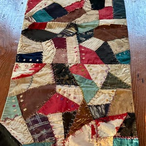 May include: A rectangular patchwork textile runner featuring an array of irregularly shaped fabric scraps in shades of red, blue, black, brown, cream, and teal. The pieces are joined together with visible decorative embroidery stitching.