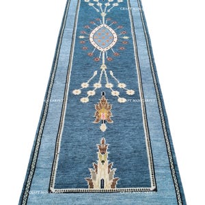Puede incluir: Una alfombra pasillera larga y rectangular en tonos azules, con un medallón central y motivos florales simétricos en crema, marrón y amarillo. La alfombra tiene un borde azul oscuro con un patrón repetitivo y flecos blancos en los extremos.