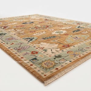 May include: A rectangular area rug featuring a golden-brown field with a repeating floral and geometric pattern in shades of sage green, red, and cream. The border is light green with intricate motifs and the text CRAFT MAN CARPET.