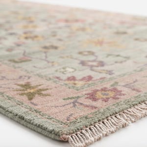 May include: A rectangular area rug featuring a light sage green background with a floral pattern in shades of pink, yellow, and dark green. The rug has a wide pink border with floral motifs and a short, light-colored fringe along the edge.