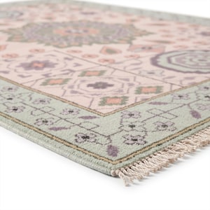 May include: A rectangular area rug featuring a pale pink center with repeating geometric and floral patterns in shades of sage green, lavender, and peach. The rug has a wide sage green border with floral motifs and short white fringe on the edge.