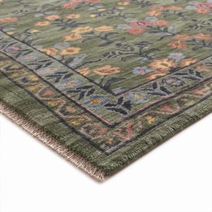 May include: A rectangular area rug featuring a deep olive green base color with a repeating floral pattern in shades of pink, yellow, and light blue. The rug includes a wide decorative border with dark blue floral motifs and a short white fringe.