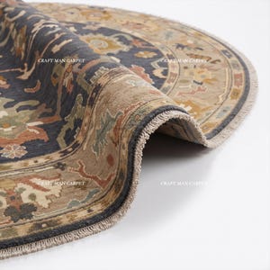 May include: A round area rug featuring a dark blue center with intricate floral patterns in tan, green, and orange. The border is a light tan with matching floral motifs and a fringed edge. The text CRAFT MAN CARPET is visible on the rug surface.