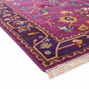 May include: A rectangular area rug featuring a vibrant magenta field with a deep purple border, decorated with multi-colored floral and geometric motifs. The rug includes white fringe at the edge and the text CRAFT MAN CARPET printed on it.