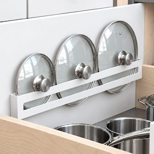 Puede incluir: Un organizador de cocina de metal blanco montado dentro de un cajón, que sostiene tres tapas de olla circulares de vidrio con pomos de metal plateado. El estante cuenta con un diseño de barra horizontal para asegurar las tapas en posición vertical.