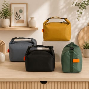 Könnte beinhalten: Vier rechteckige Lunchtaschen mit Rollverschluss und schwarzen Kunststoffschnallen. Die Taschen sind in Senfgelb, Anthrazit, Schwarz und Waldgrün erhältlich. Jede Tasche verfügt über eine kleine, leuchtend orangefarbene Stofflasche an der Seite.