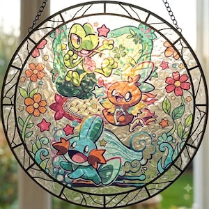 Pode incluir: Um apanhador de sol circular em vitral com três criaturas coloridas de desenho animado rodeadas por flores, folhas e pequenas estrelas. O design inclui uma personagem verde, uma criatura laranja e uma criatura aquática azul.