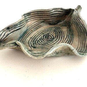 May include: A hand-formed ceramic dish with an irregular, wavy edge and a leaf-like shape. The surface features a light gray and off-white glaze with dark, etched concentric circular lines that mimic the appearance of wood grain patterns.