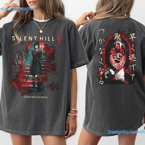 May include: A charcoal grey t-shirt featuring a Silent Hill f graphic on the front with a character in a school uniform and the text Friends don't last forever. The back displays a red and black abstract face graphic with Japanese text.