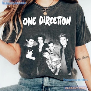 May include: A charcoal gray short-sleeve t-shirt featuring the white text ONE DIRECTION at the top. Below the text is a black and white photographic print of the four members of the band One Direction posing together in a group portrait.