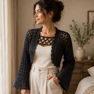 May include: A dark charcoal gray open-front crochet bolero featuring long bell sleeves, a scalloped edge trim, and an intricate lattice pattern across the upper chest area, worn over a plain cream-colored tank top and light beige trousers.