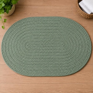 Könnte beinhalten: Ein ovaler Teppich aus salbeigrünem Garn in einem durchgehenden, konzentrischen Häkelstichmuster. Der Teppich weist eine dichte, strukturierte Oberfläche auf, die durch die sich wiederholenden Garnschleifen entsteht und eine einheitliche grüne Farbe bildet.