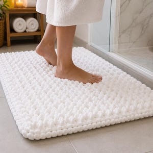 May include: A rectangular white bath mat featuring a chunky, hand-knitted texture with thick, soft yarn loops. The mat has a dense, cushioned appearance and is positioned on a tiled floor, providing a plush surface for bare feet after showering.