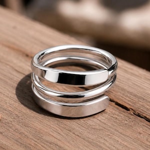 May include: A polished silver-tone ring featuring a multi-layered spiral design. The band consists of three distinct, smooth, and reflective coils that wrap around to create a continuous, stacked appearance with rounded edges and a high-shine finish.