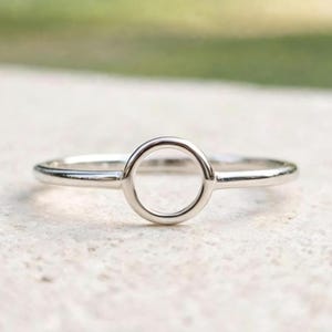 Puede incluir: Un anillo de tono plateado pulido que presenta una banda delgada y lisa con un diseño central de aro circular abierto. El metal tiene un acabado metálico reflectante que capta la luz, creando una estética limpia y sencilla para esta pieza de joyería.