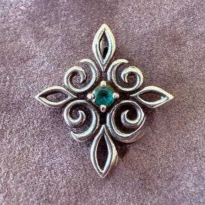 May include: A silver pendant featuring a symmetrical, openwork scroll design arranged in a diamond shape. At the center of the metalwork is a single, round, faceted green gemstone held in a four-prong setting, with a bail at the top.