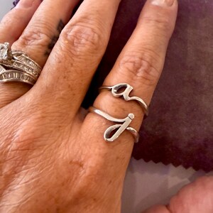 Op de afbeelding: Twee zilverkleurige ringen gedragen aan een vinger, elk voorzien van een enkele kleine cursieve letter. De ene ring toont de letter a en de andere ring toont de letter j. Een aparte zilveren ring met meerdere stenen is zichtbaar aan een vinger ernaast.