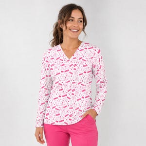 May include: A long-sleeve V-neck top featuring a white base with a repeating pink pattern of nurse caps, syringes, hearts, and the text "NURSE," "RN," "CARE," and "XOXO." The top is paired with solid pink pants, creating a coordinated set.