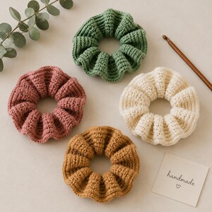 May include: Four circular crochet hair scrunchies in sage green, dusty rose, cream, and mustard yellow. A brown wooden crochet hook is positioned near the cream scrunchie. A small white square card features the printed text handmade and a heart symbol.