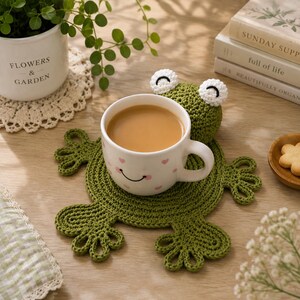 May include: A round, olive green crocheted frog coaster with four webbed feet and two white, circular eyes with black stitching. A white ceramic mug with a small pink heart pattern and a simple black line smile sits on the coaster.