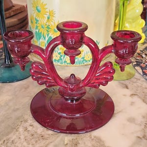 Puede incluir: Un candelabro de cristal rojo rubí con tres portavelas dispuestos en fila horizontal. El soporte central está elevado sobre los dos laterales, que están flanqueados por elementos decorativos de cristal en forma de abanico sobre una base circular.