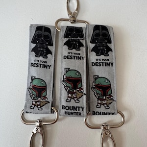 May include: Three grey fabric key fobs featuring cartoon illustrations of Darth Vader with the text IT'S YOUR DESTINY and Boba Fett with the text BOUNTY HUNTER. Each strap includes silver-toned metal swivel lobster clasps at both ends.