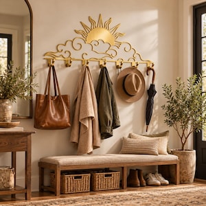 May include: A gold-toned metal wall-mounted coat rack featuring a decorative sun and cloud design. The rack includes eight hooks holding a brown leather tote bag, a beige blanket, a green jacket, a tan fedora hat, and a black umbrella.