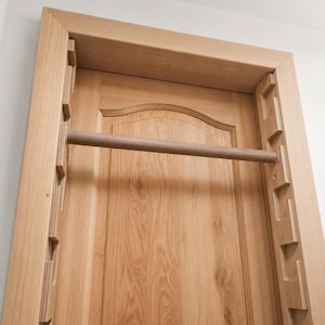 Può includere: Una barra per trazioni in legno marrone chiaro montata all'interno di un telaio di porta. Il sistema presenta due pannelli laterali verticali con molteplici fessure per la regolazione dell'altezza, che sostengono una barra cilindrica.