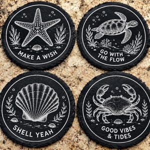 May include: A set of four round black slate coasters featuring white engraved marine life illustrations. The designs include a starfish with the text MAKE A WISH, a sea turtle with GO WITH THE FLOW, a seashell with SHELL YEAH, and a crab with GOOD VIBES & TIDES.