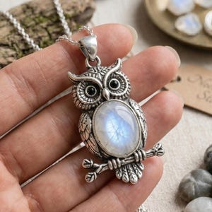 Peut inclure: Un collier pendentif hibou argenté avec une grande pierre de lune ovale irisée sertie au centre du corps. Le hibou présente des plumes en métal détaillées, de petits yeux noirs et est perché sur une base en métal en forme de branche.