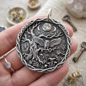 Puede incluir: Un colgante circular de plata con un relieve detallado de un búho posado en una rama de árbol sosteniendo una llave. El diseño incluye una luna, estrellas, runas, helechos y setas. El búho tiene ojos de piedra azul y el colgante está enmarcado por un borde de vid trenzada con una anilla superior.