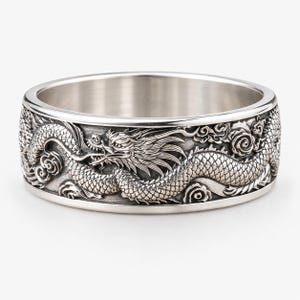 Puede incluir: Un anillo de metal plateado con un diseño de dragón en relieve detallado que envuelve toda la banda exterior. El dragón está representado con escamas intrincadas, una melena fluida y motivos de nubes estilizados sobre un fondo oscuro.