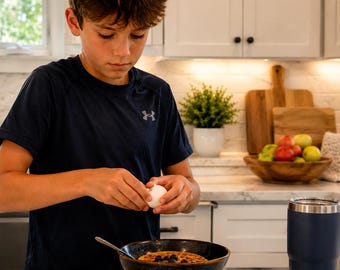 May include: A dark navy blue short-sleeved athletic shirt with a small grey logo on the chest. The shirt is worn while cracking a white egg over a dark speckled bowl containing a waffle topped with blueberries, next to a dark blue YETI tumbler.