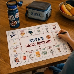 May include: A white rectangular paper chart titled KUYA'S DAILY ROUTINE with a grid of illustrated tasks including a start flag, breakfast, reading, vitamins, jersey, toothbrush, shower, clothes, apple, piggy bank, lunch, map, soccer ball, and calculator.